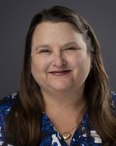 Middle aged caucasian woman with long brown hair with a slight smile, wearing a blue and white blouse and a gold locket.