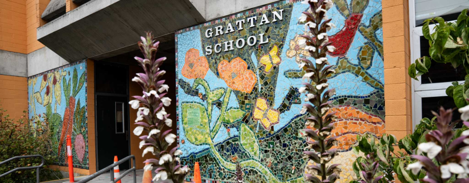 An outdoor wide shot shows a colorful mosaic mural on the exterior wall of Grattan School in San Francisco. The mural features vibrant tilework depicting orange flowers, yellow butterflies, and a red bird against a blue background. The words "GRATTAN SCHOOL" are displayed in white capital letters within the mosaic. In the foreground, two tall, purple and white flower stalks are slightly out of focus, framing the view of the school's entrance. Photo Credit: Anna Connors / S.F. Chronicle