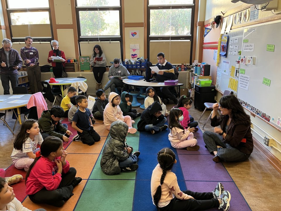 Lower grade students gathered on rug for lesson. Adults observing teacher. 