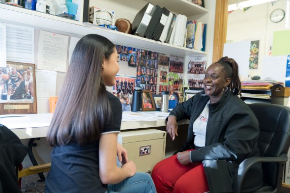 Rear view of a woman with long dark hair sitting across from an african american woman who has on red pants, a white tee shirt and a black jacket, her hair is in a pony tail and she is smiling.