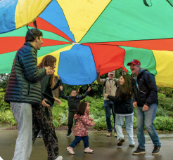 A group of 7 adults and children under a bright multi colored parachute