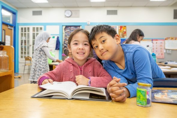two students sit at a desk, heads together, looking at the camera