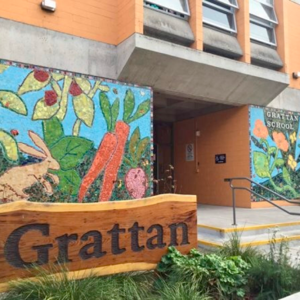 A vibrant outdoor shot of the entrance to Grattan School, featuring a large, natural wood sign in the foreground with the word "Grattan" carved in dark letters.  Behind the sign, the school's facade is decorated with colorful mosaic murals depicting a nature scene with a rabbit, large orange carrots, and green leaves against a bright blue background. To the right, a set of concrete stairs with a metal handrail leads to the recessed entrance, next to which is another mosaic panel and a sign reading "GRATTAN"
