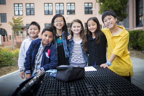A group of 6 middle school students stands around a table in front of a school
