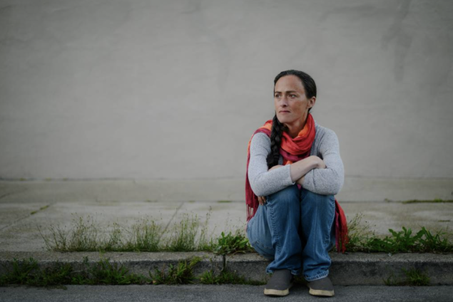 Woman with brown hair sitting on the sidewalk with her arms folded over her knees