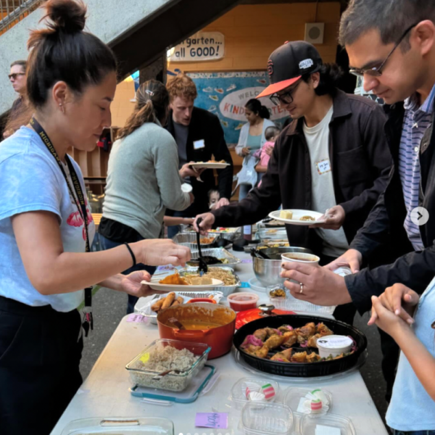An image showing a long buffet table covered with a variety of aluminum trays and colorful dishes of food during an Affinity Night at Grattan Elementary. Several adults are standing around the table, serving themselves from dishes that include rice, spring rolls, and various stews.
