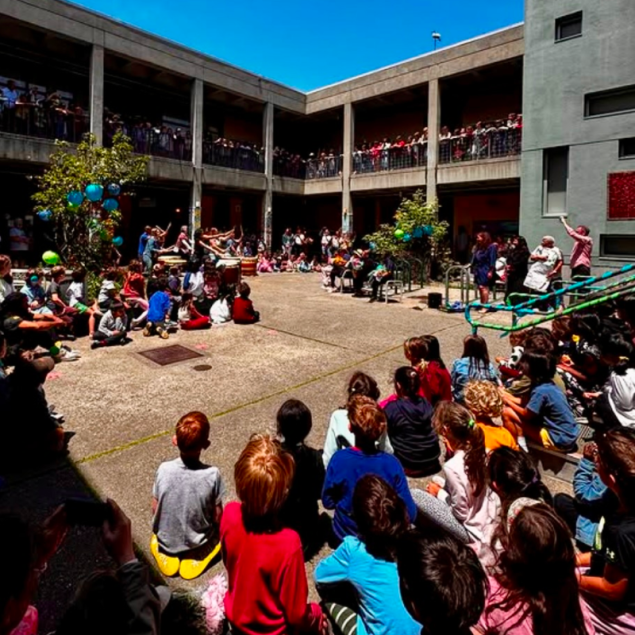 An image showing the Grattan Elementary courtyard during a Clap Out ceremony. A large crowd of students and staff is gathered along the upper balconies of the multi-story school courtyard. In the center of the sunny courtyard, a teacher-led taiko drumline celebrates the retiring honorees.