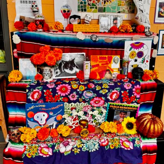 An image showing a vibrant Day of the Dead altar, or ofrenda, decorated with bright orange marigolds, traditional papel picado banners, and framed photographs of loved ones. The tiered display is set up in a school hallway, featuring colorful sugar skulls and candles as part of the community-led cultural celebration.