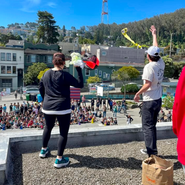 An image showing two adults on the flat, gravel-covered roof of Grattan Elementary School in San Francisco, preparing to drop student-designed egg-protection inventions to a large crowd of students gathered in the courtyard below. One person is shown mid-toss, releasing a yellow device with a long streamer, while the other holds a red and black parachute-style contraption. Sutro Tower and the houses of Cole Valley are visible in the background under a clear blue sky.