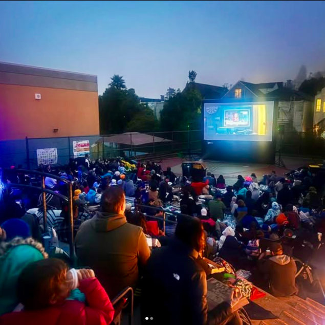 An image showing a large group of families and community members gathered in the Grattan elementary courtyard at dusk for an outdoor movie night. Attendees are seated on blankets and low chairs across the yard, facing a large inflatable projector screen that displays a brightly lit animated scene. The surrounding neighborhood houses and trees are visible against an evening sky.