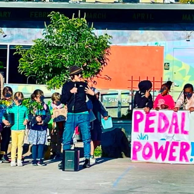 A vibrant outdoor scene at Grattan Elementary featuring a group of elementary students and an adult speaker on a paved school yard. The speaker, wearing a black sweatshirt, hat, and sunglasses, holds a microphone while gesturing to the group. Several students stand nearby, some holding potted plants, while others stand behind a large hand-painted sign that reads "PEDAL POWER!" in bright pink letters. The background shows a school building with windows and a colorful mural.