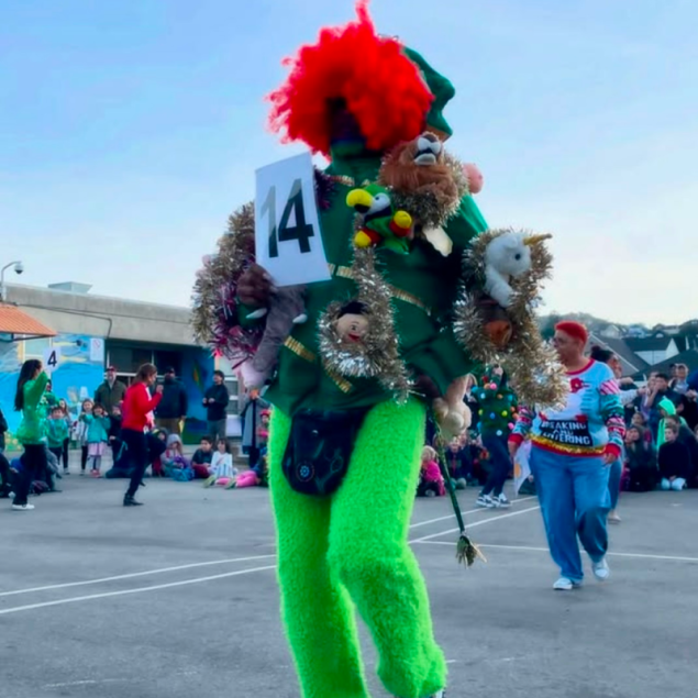 An image showing a person in an elaborate costume during a Spirit Week event at Grattan Elementary. The person is wearing bright neon green fuzzy pants, a red afro wig, and a green top heavily decorated with tinsel and numerous stuffed animals, while holding a sign with the number 14. In the background, other community members and students are gathered in the sunny school yard under a clear sky.