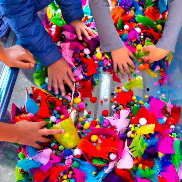 An image showing a bird's-eye view of a clear sensory bin filled with vibrant craft materials. Multiple pairs of children's hands reach into the bin, which is overflowing with bright feathers, colorful pom-poms, and small plastic cups. The assortment includes a mix of textures and bold primary colors like red, blue, yellow, and neon pink.