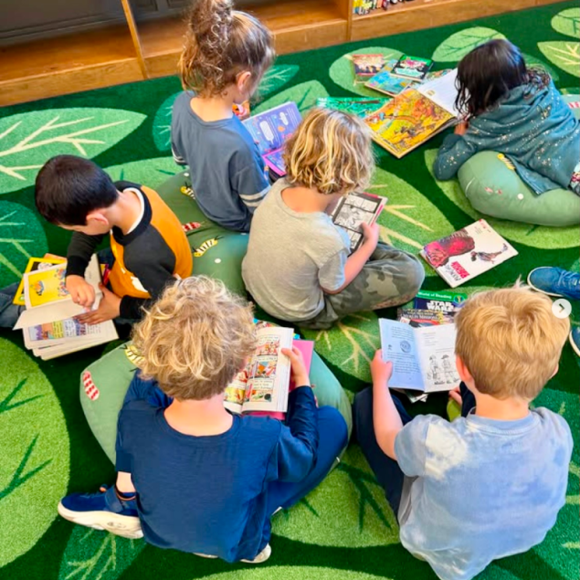 An image showing a top-down view of six elementary students sitting in a circle on a vibrant green rug decorated with large leaf patterns. The students are deeply engaged in reading various books, including graphic novels and picture books, which they have likely selected during the Book Swap event. Several other books are scattered on the rug around them, and some children are resting on large, leaf-shaped floor cushions.