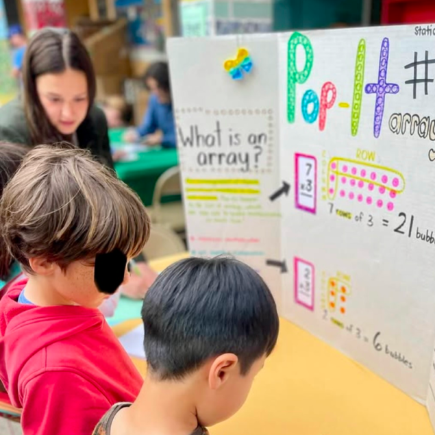 An image showing two elementary-aged students and an adult mentor looking closely at a handmade educational display board at Grattan's Math Night. The colorful board, titled "Pop-It Arrays," features the question "What is an array?" and uses diagrams of bubble-pop toys to illustrate multiplication concepts like $7 \times 3 = 21$. In the background, other community members are engaged at a table covered with a green cloth.