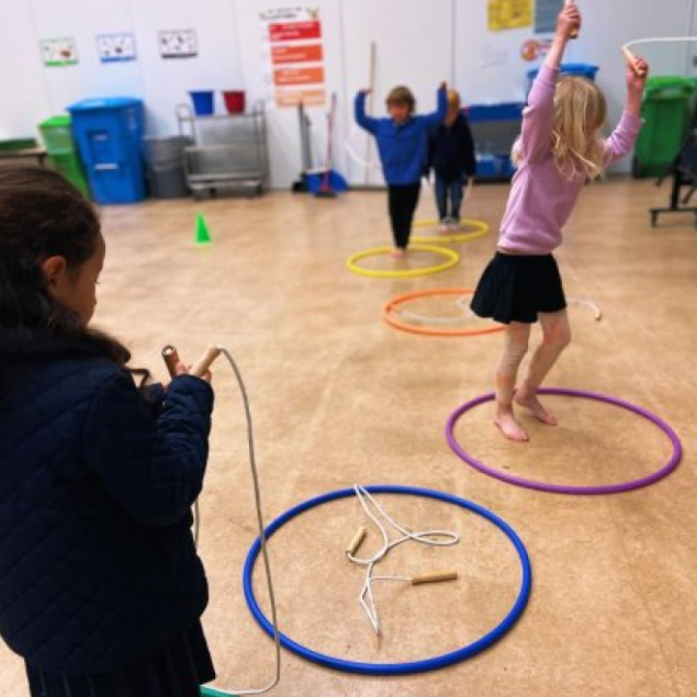 An image showing several elementary students participating in a Physical Education (PE) activity in an indoor gym or multipurpose room. In the foreground, a student in a dark blue quilted jacket holds a jump rope, looking toward other children who are jumping in and out of colorful hula hoops—blue, purple, orange, and yellow—laid out in a line on the tan floor. One student in a pink sweater and black skirt is captured mid-jump inside a purple hoop, while others in the background follow along with their arms