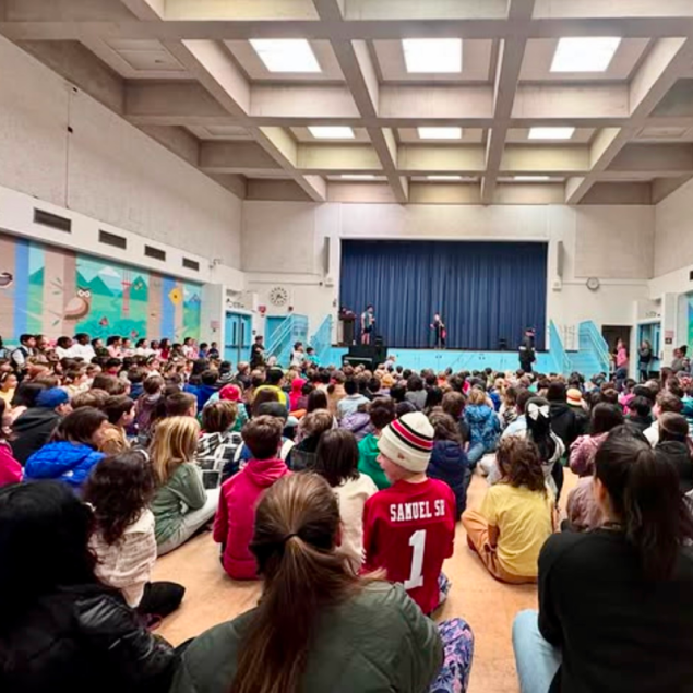 An image showing a large audience of elementary students and adults sitting on the floor of a high-ceilinged auditorium, watching a talent show performance. Two students are on a raised stage in front of a blue curtain, with one standing at a podium and the other performing in the center. The room features a distinctive coffered ceiling with skylights and a large colorful mural on the left wall depicting an owl and San Francisco landmarks.