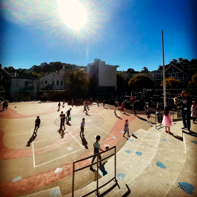 An image showing a sunny outdoor school yard where numerous students are actively playing with balls and hula hoops. The ground is painted with large tan and reddish-brown curved patterns and white game lines. In the lower right, several rows of concrete bleacher-style steps lead down to the yard, with a few children and an adult standing nearby. The background features neighborhood houses and lush green trees of Cole Valley under a bright, clear blue sky with a prominent sun.