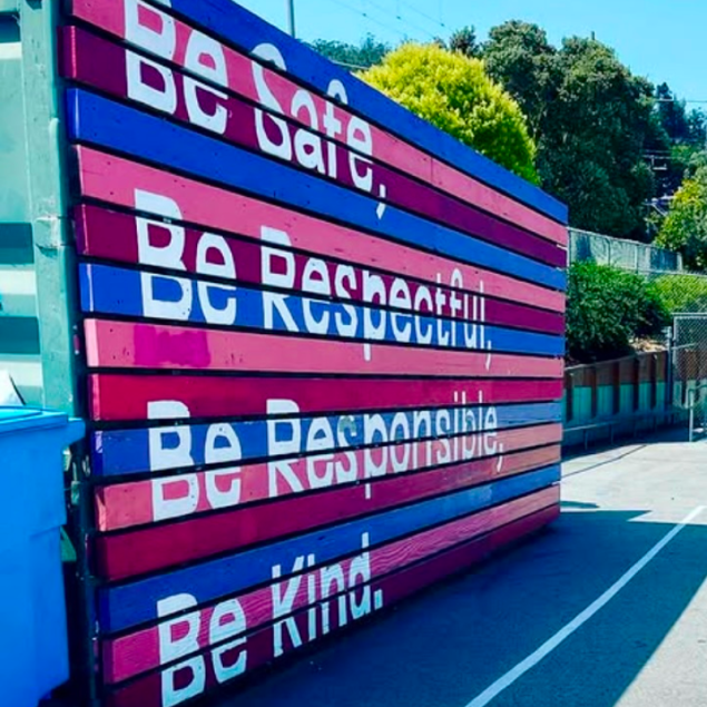 The image shows a large, vibrant mural painted on a slatted wooden fence or container wall in the Grattan Elementary schoolyard. The mural consists of horizontal slats painted in alternating shades of bright pink, magenta, and deep blue.  The core tenets of "The Grattan Way" are painted in large, white, sans-serif block lettering across the slats:  Be Safe,  Be Respectful,  Be Responsible,  Be Kind.