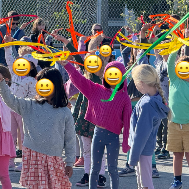 An image showing a group of students in an outdoor school yard participating in a dance or movement performance for the VAPA Showcase. Several children are raising their arms to wave long, vibrant ribbons in shades of red, yellow, and orange. The students are dressed in colorful everyday clothing
