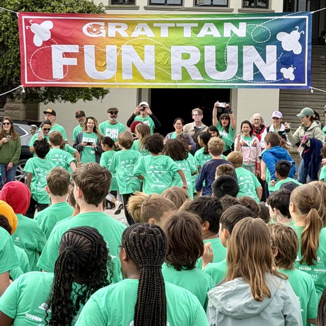 A large crowd of children in matching teal t-shirts gathers excitedly beneath a vibrant rainbow banner that reads "GRATTAN FUN RUN."